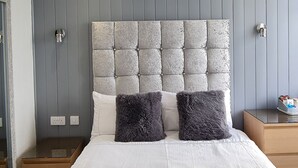 A bedroom with a tufted headboard, a large bed with white linens, two dark gray pillows, and a gray throw blanket. - Ocean Bay Hotel (Blackpool)