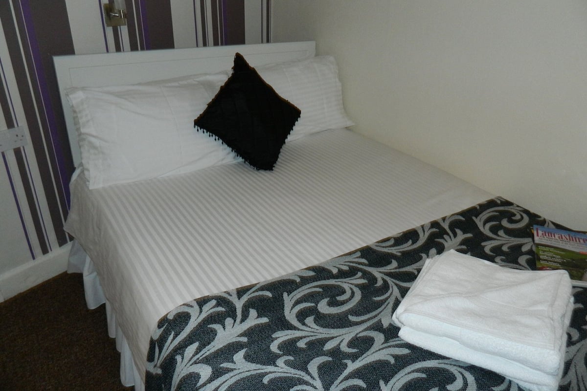 A single bed with white bedding and a black pillow, a patterned blanket, and a folded white towel on the bed.
