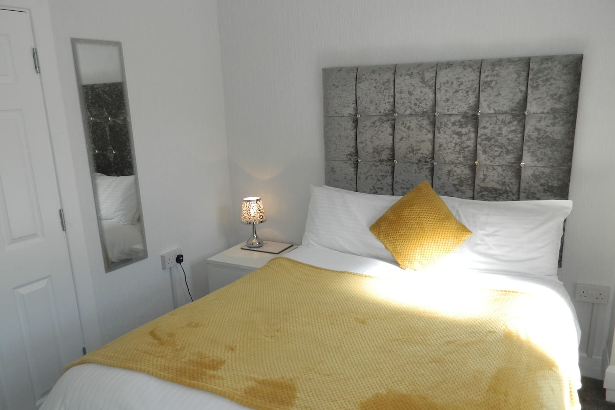 A neatly made bed with a white and yellow bedspread, a grey tufted headboard, and a bedside lamp.