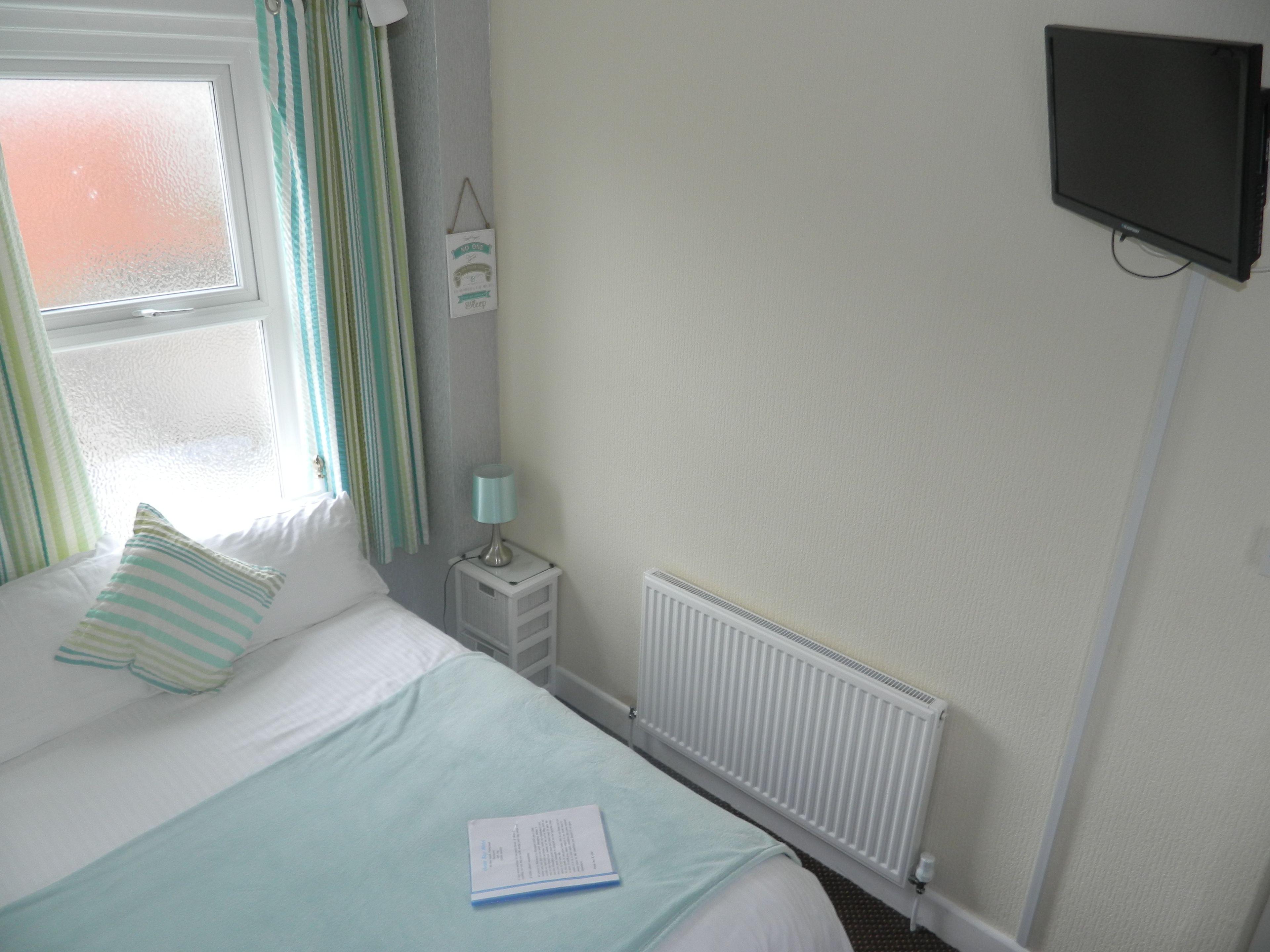 A neatly made bed with a striped pillow, a bedside table with a lamp, a wall-mounted TV, and a window with curtains.