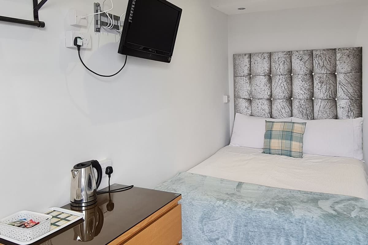 A modern bedroom with a bed, a bedside table with a kettle, a flat-screen TV mounted on the wall, and a wooden dresser.