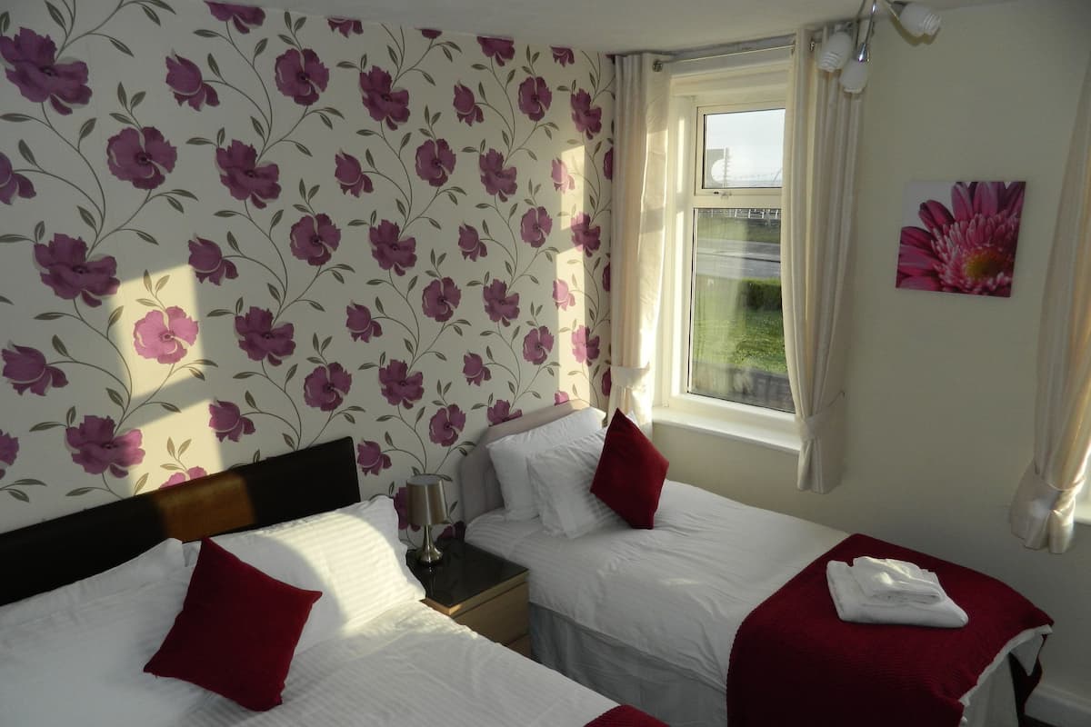 A hotel room with two beds, floral wallpaper, a window with curtains, and a painting on the wall.