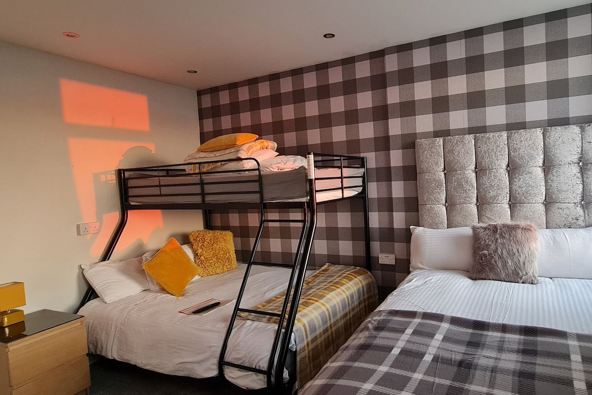 A hotel room with a bunk bed, a single bed, a checkered wall, a headboard, and a bedside table with a lamp.