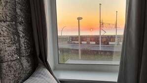 A bedroom with a large window, a view of a sunset, and a fur throw on the bed. - Ocean Bay Hotel (Blackpool)