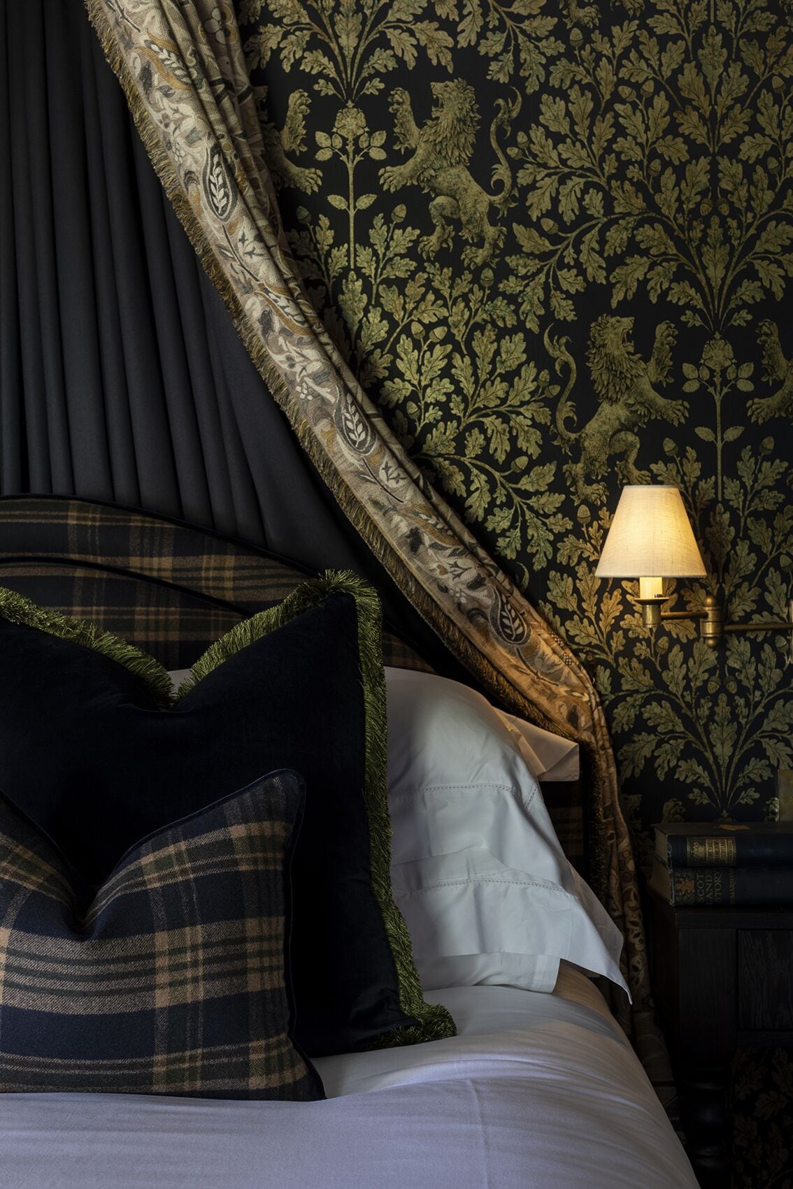 Una cama con papel tapiz verde oscuro y detalles dorados, una lámpara y almohadas a cuadros.