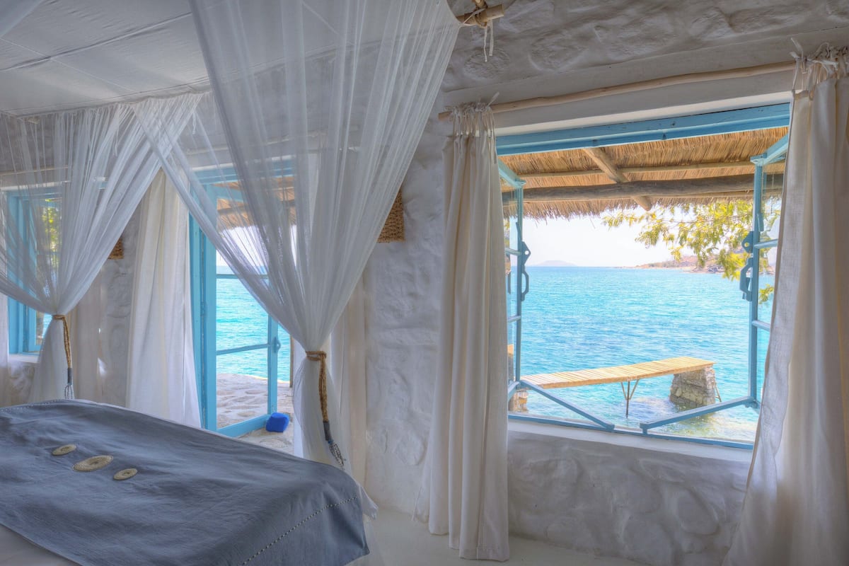 A bedroom with a bed, a window with a view of the sea, and a thatched roof.