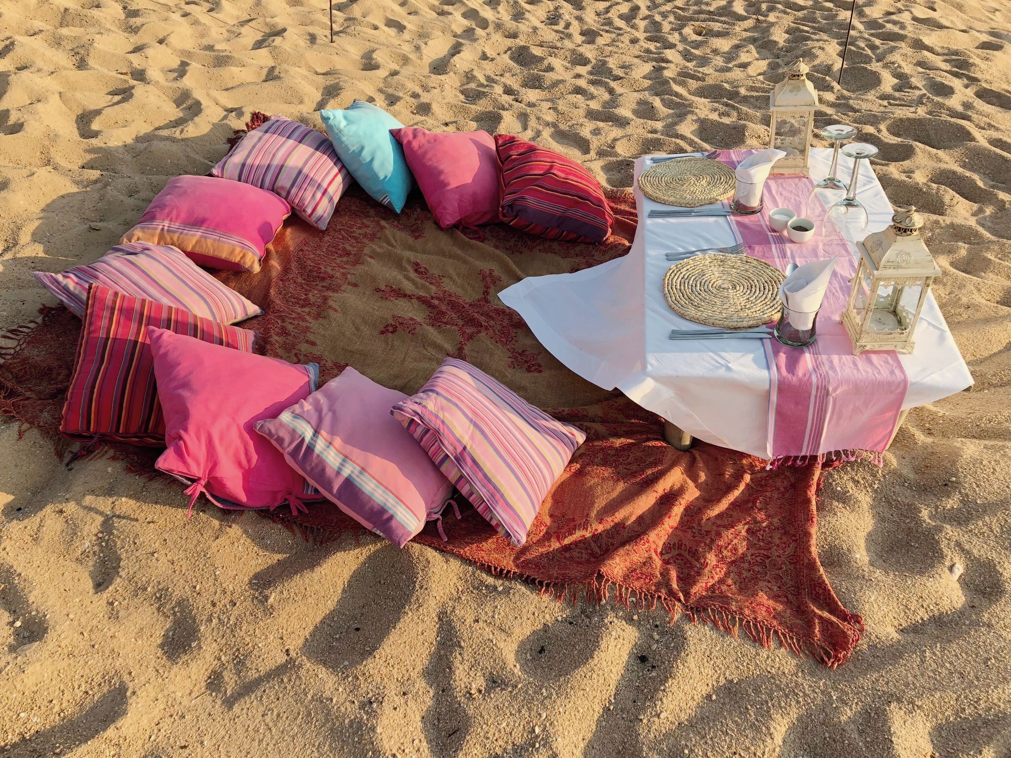A circular seating arrangement with colorful cushions and a table set for a meal on a sandy beach.