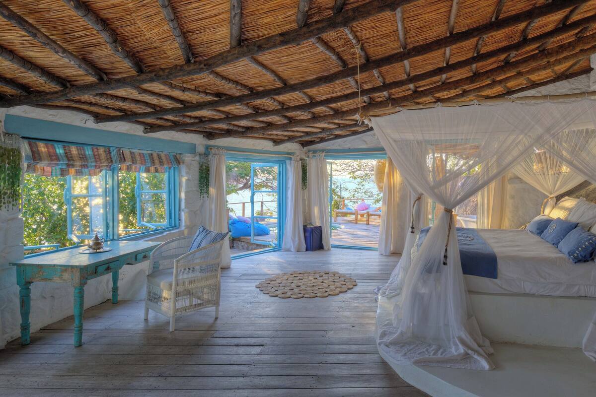 A cozy room with a wooden floor, a bed with white canopy, and a window with a view of greenery.