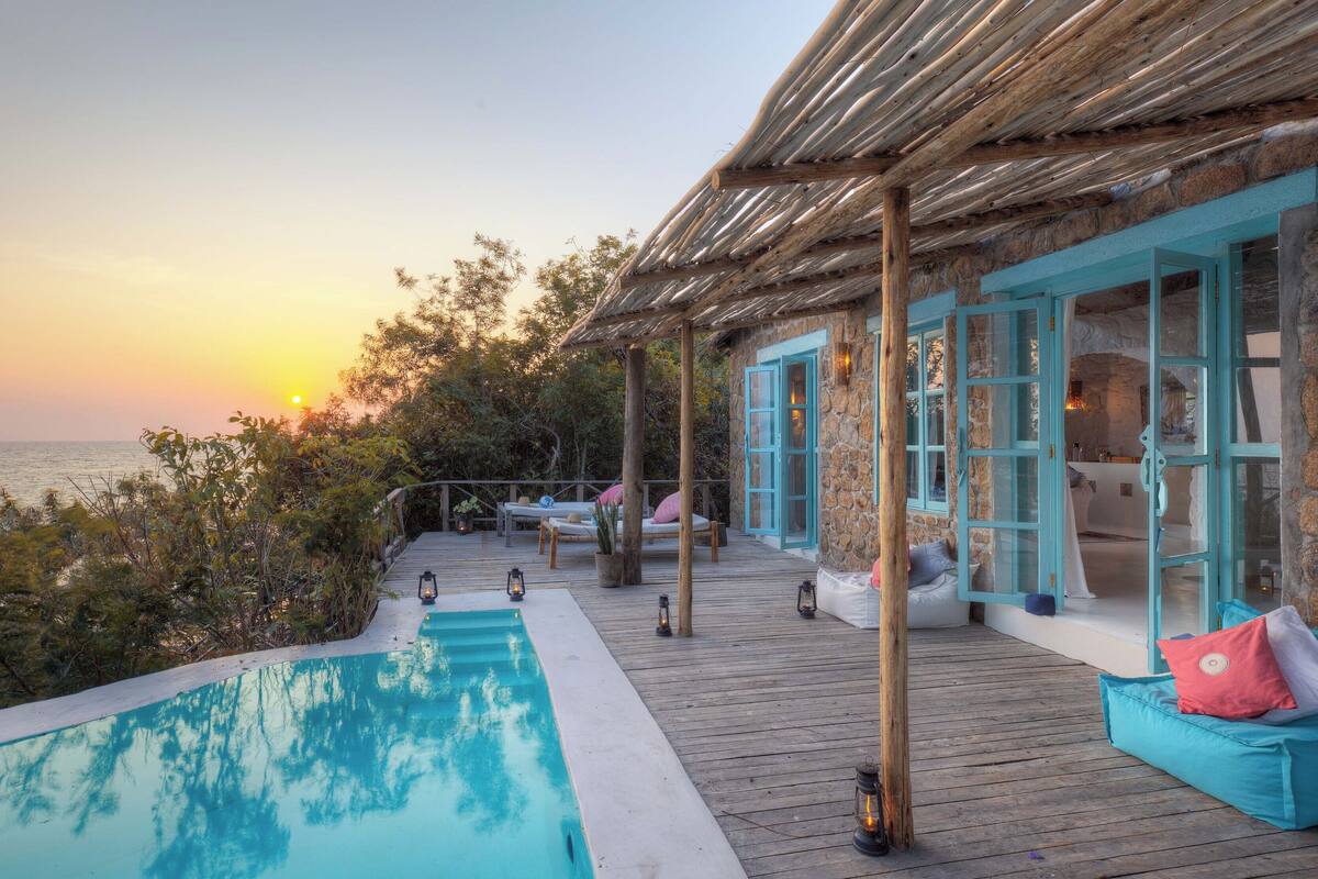 A wooden deck with a pool, stone walls, and a view of the sea at sunset.