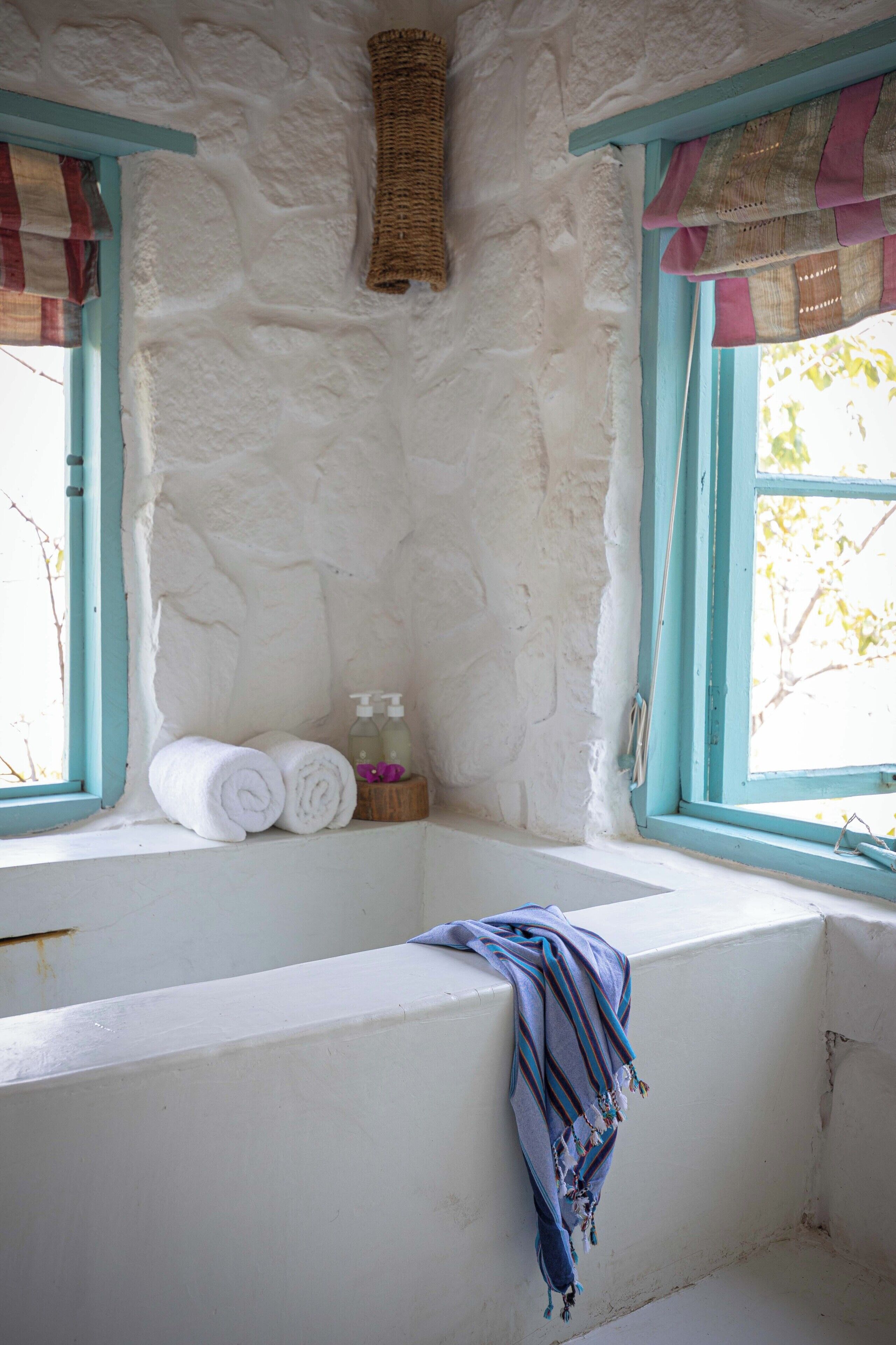 A bathroom with a stone wall, a window with blue trim, and a towel rack with rolled towels and toiletries.