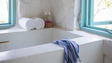 A bathroom with a stone wall, a window with blue trim, and a towel rack with rolled towels and toiletries.