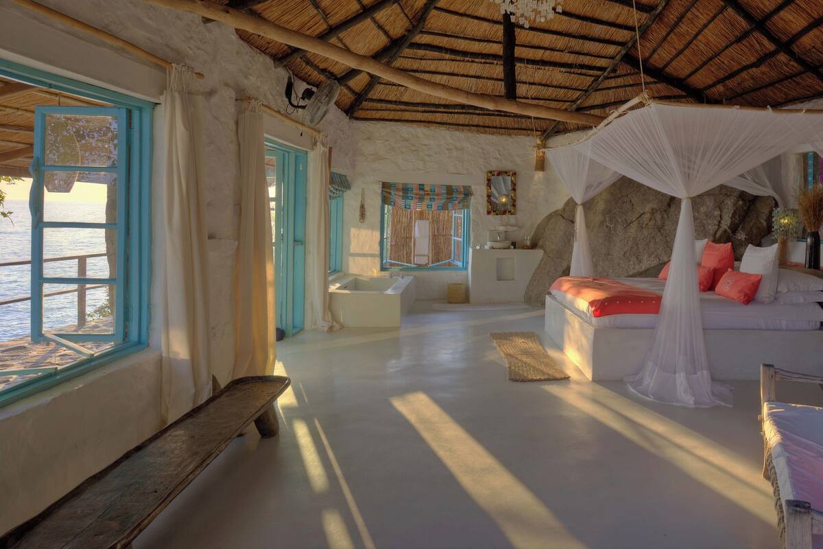 A room with a bed, a bench, a bathtub, and a view of the sea.
