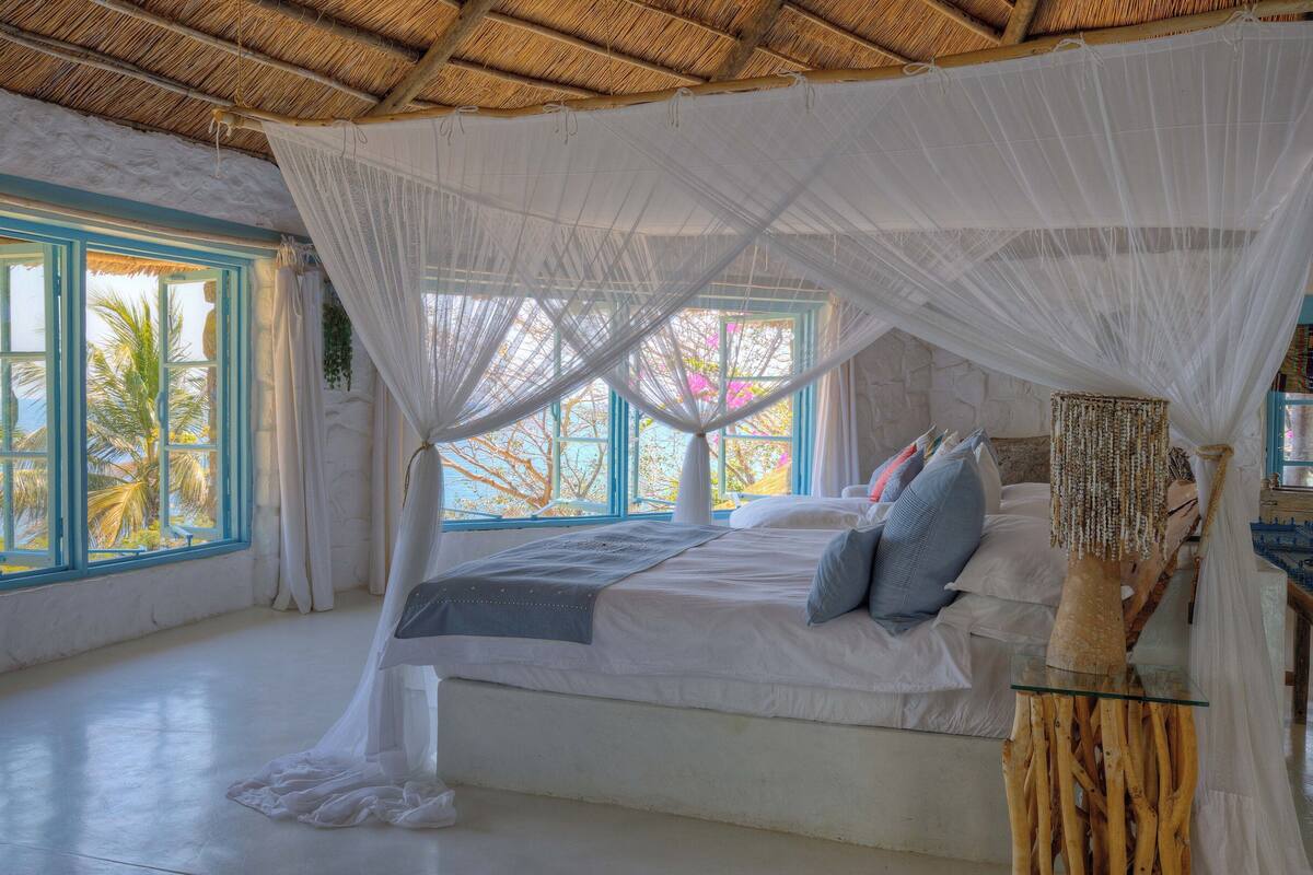 A spacious bedroom with a large bed, a canopy, and a view of palm trees through the windows.