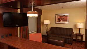 A hotel room with a wooden desk, a flat-screen TV, a sofa, two lamps, and a framed picture on the wall.