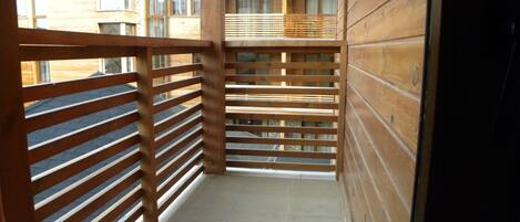 A narrow balcony with wooden railings, a tiled floor, and a view of a multi-story building with windows.