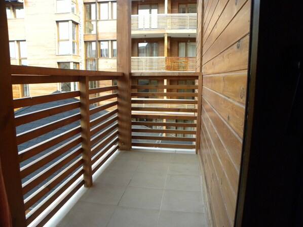 A narrow balcony with wooden railings, a tiled floor, and a view of a multi-story building with windows.