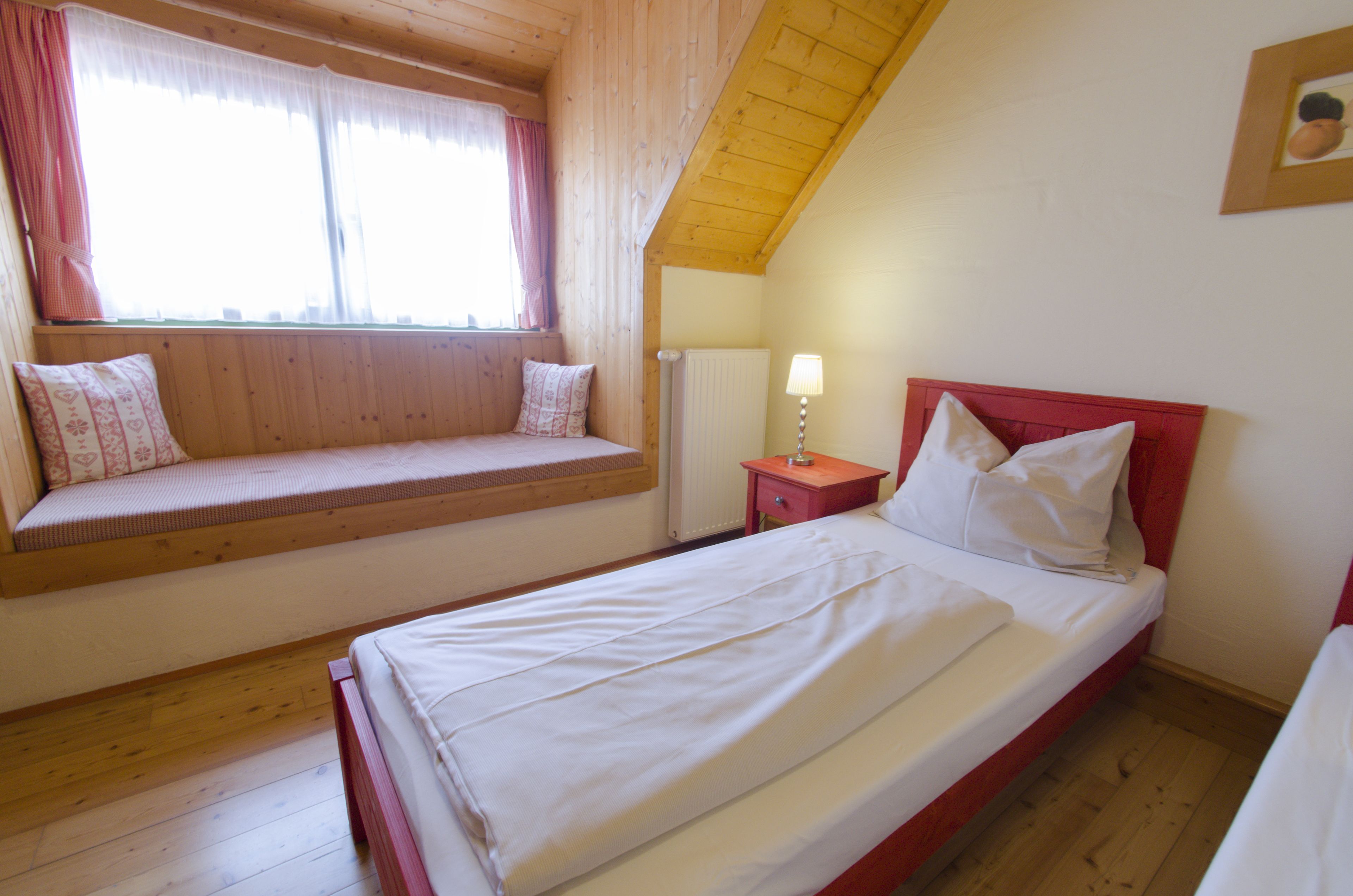 A bedroom with a wooden sloped ceiling, a window with curtains, a bedside table with a lamp, and a bed with white bedding.