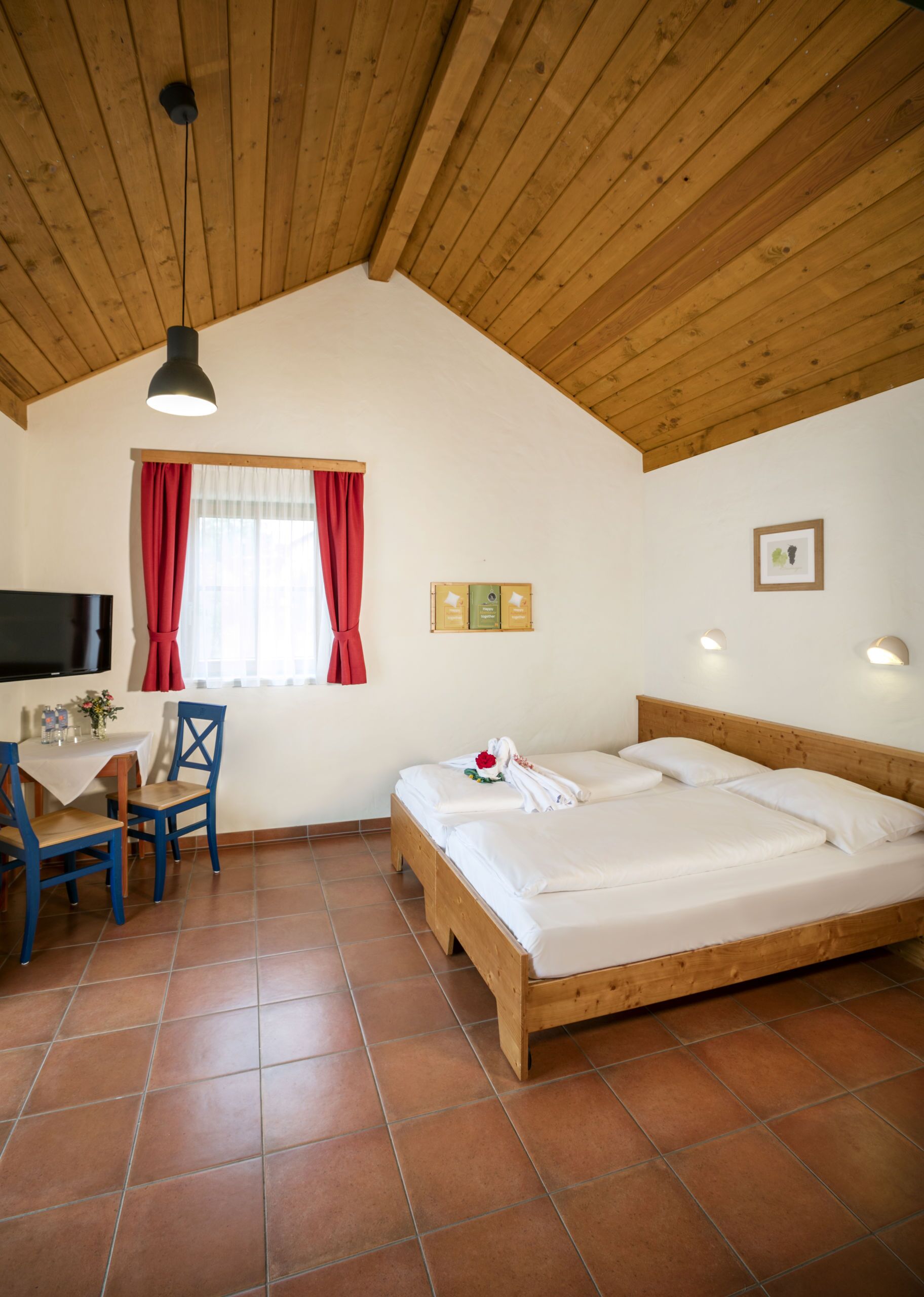 Una stanza con soffitto in legno, un letto, una scrivania con una sedia, una TV e una finestra con tende rosse.