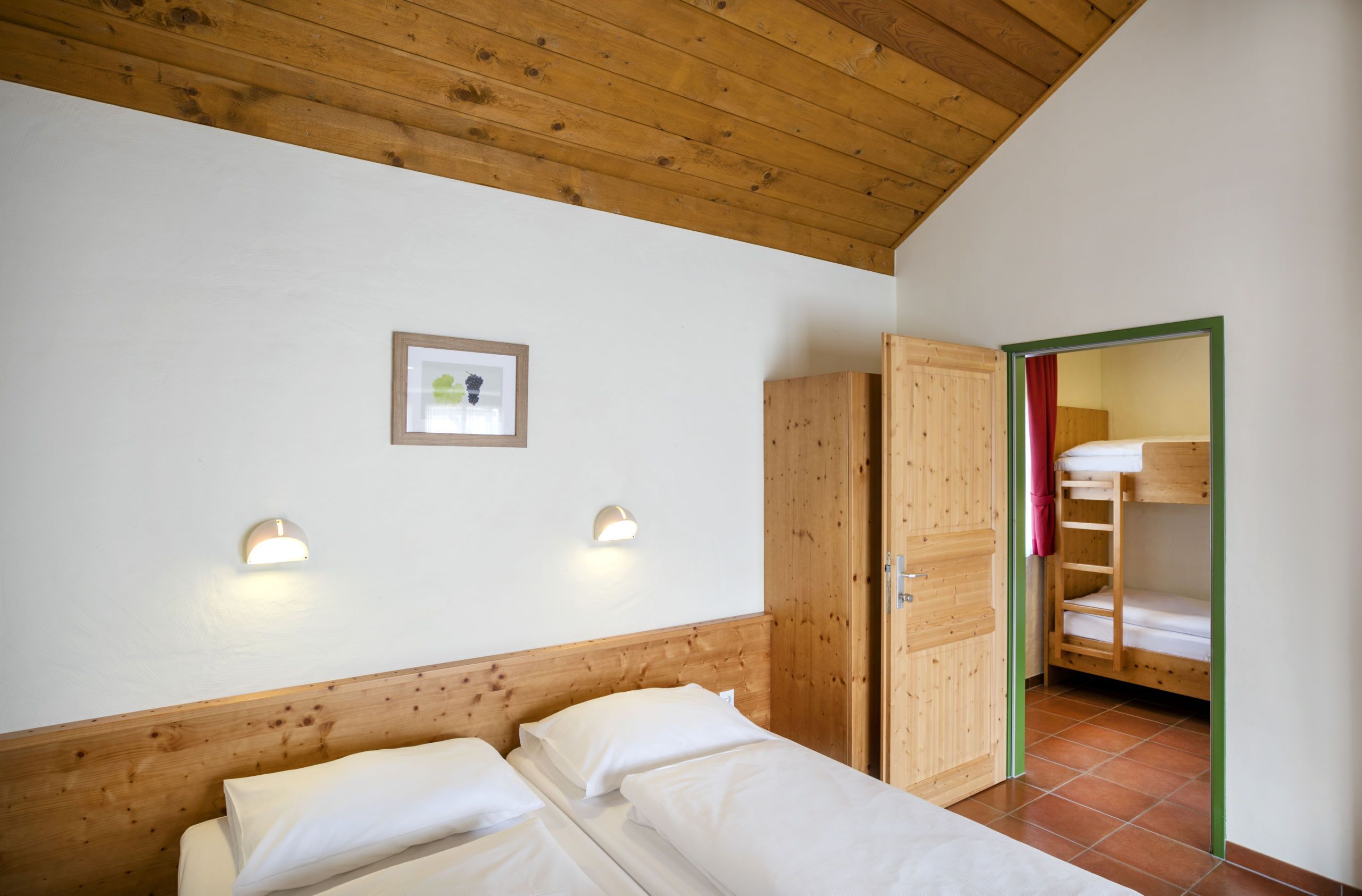 Un dormitorio con cabecera de madera, dos lámparas de noche, un cuadro enmarcado en la pared y una cama litera en la habitación contigua.
