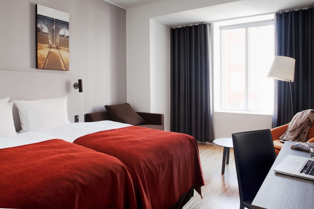 A hotel room with two beds, a desk with a laptop, a chair, a small table, and a window with curtains.