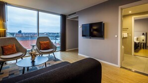 A modern hotel room with a large window offering a city view, a flat-screen TV mounted on the wall, and a cozy seating area with two armchairs and a sofa.