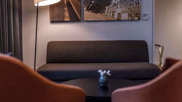 Una habitación de hotel moderna con un sofá gris oscuro, dos sillones marrones, una mesita redonda y una lámpara de pie. Dos fotografías enmarcadas decoran la pared.