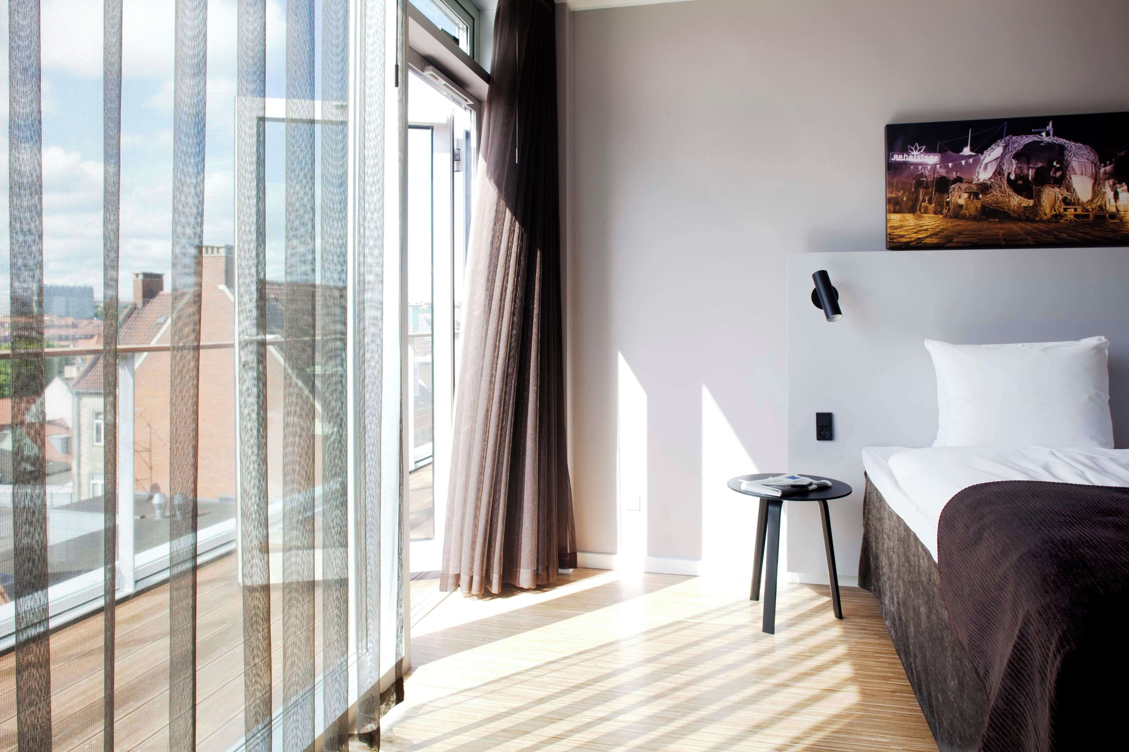 Une chambre d’hôtel avec une grande fenêtre, un lit, une petite table et un tableau au mur.
