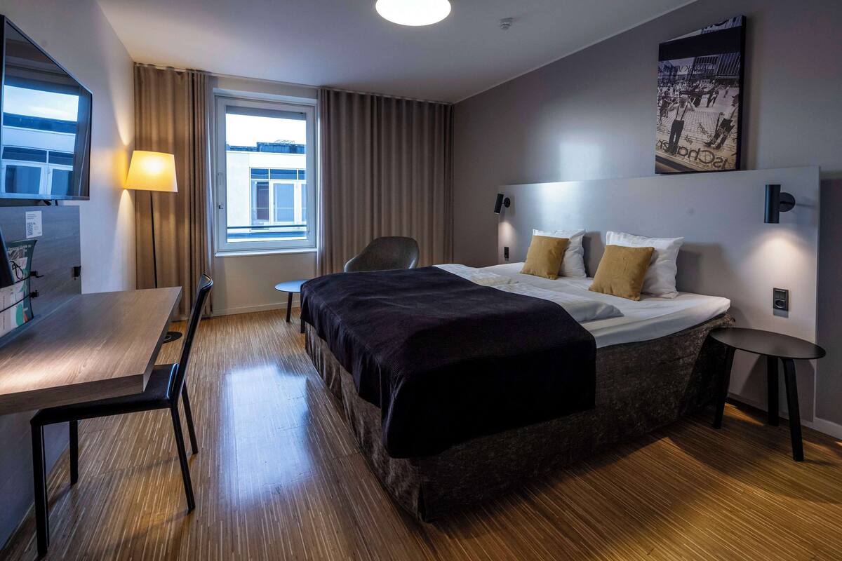 A hotel room with a bed, a desk, a chair, a small table, a lamp, and a window with curtains.