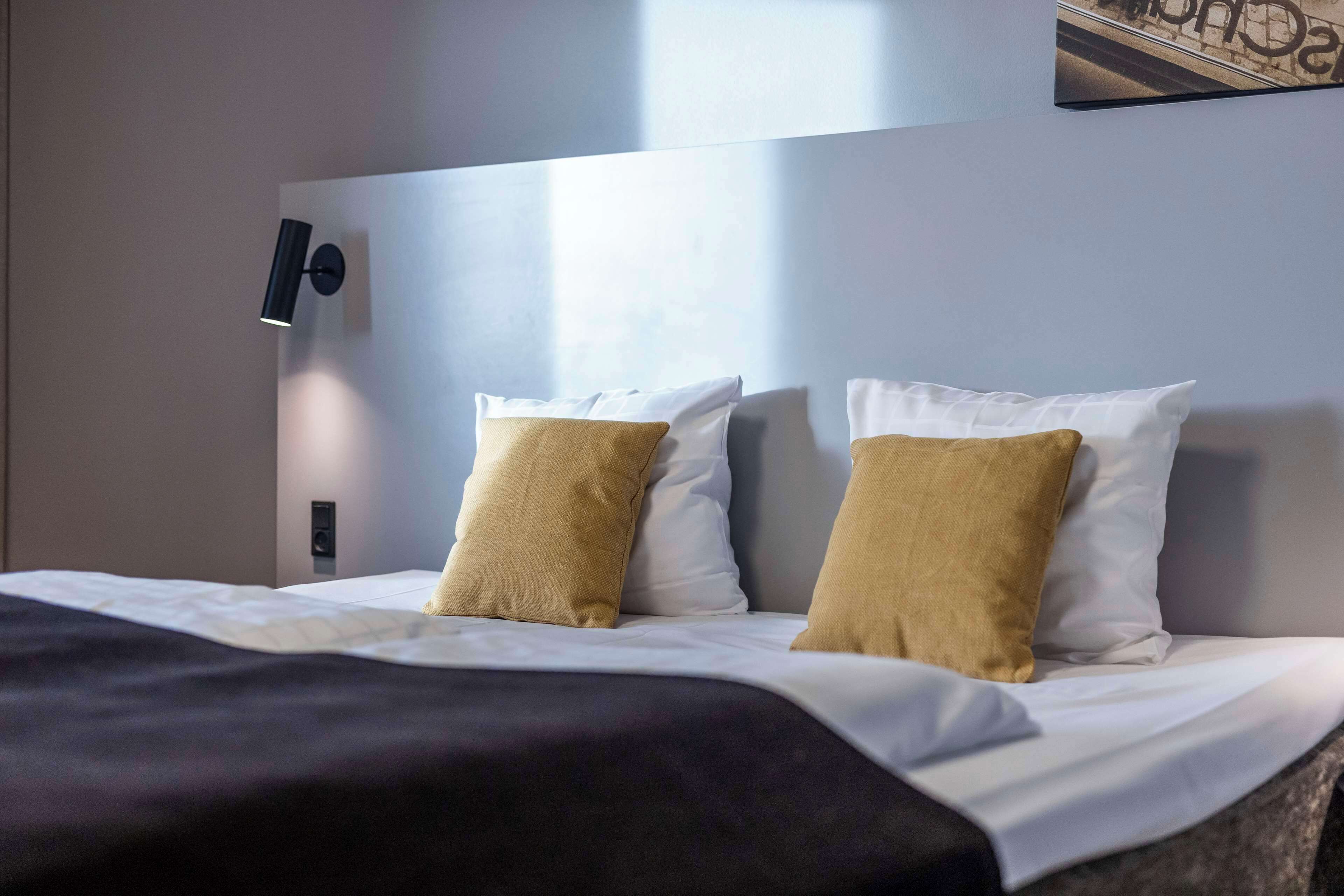 A neatly made bed with white and mustard-colored pillows, a black headboard, and a wall-mounted light fixture.
