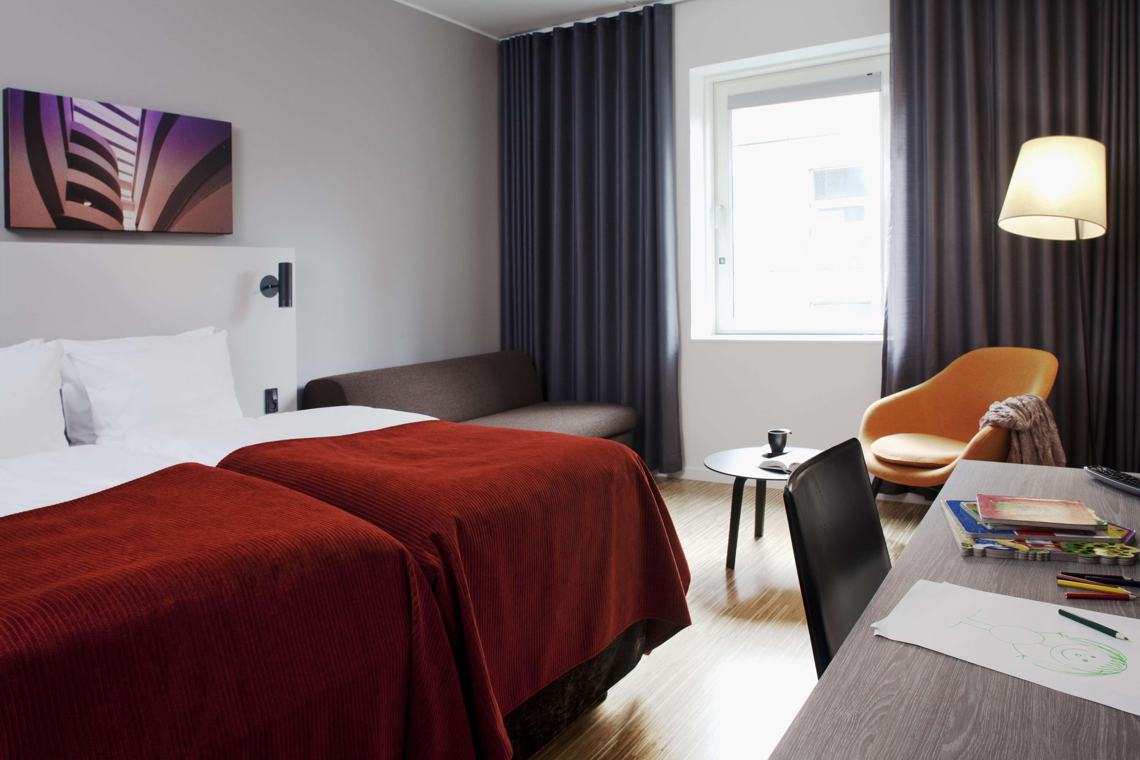 Una habitación de hotel con una cama, un escritorio con libros y una lámpara, una silla y una ventana con cortinas.
