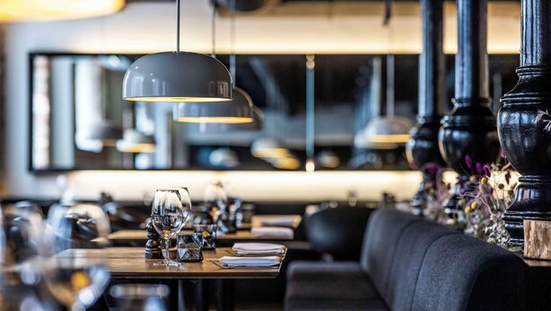A restaurant interior with black upholstered seating, tables set with wine glasses, and pendant lighting.