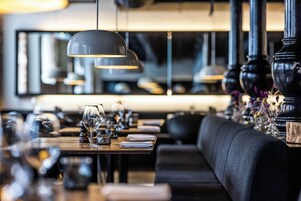 En restaurantindretning med polstrede sorte siddepladser, borde dækket med vinglas og hængende lamper.