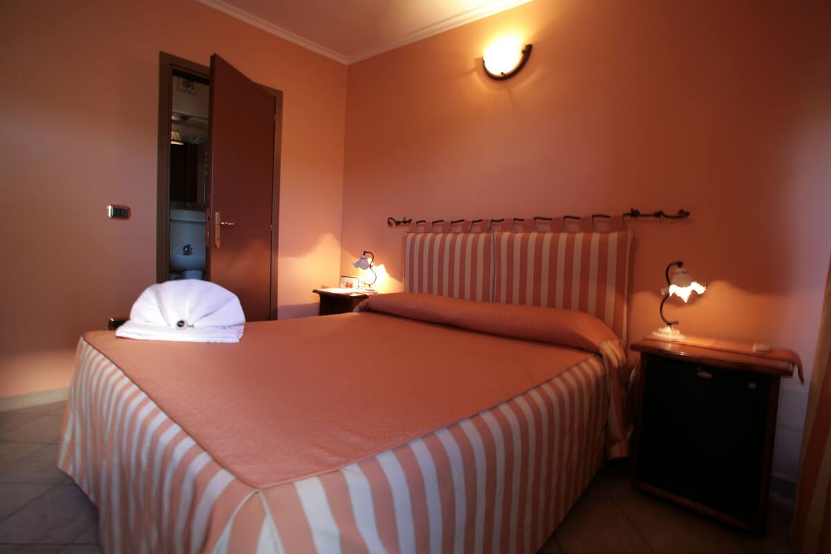 A hotel room with a bed, a nightstand, a lamp, a towel, and a door to the bathroom.