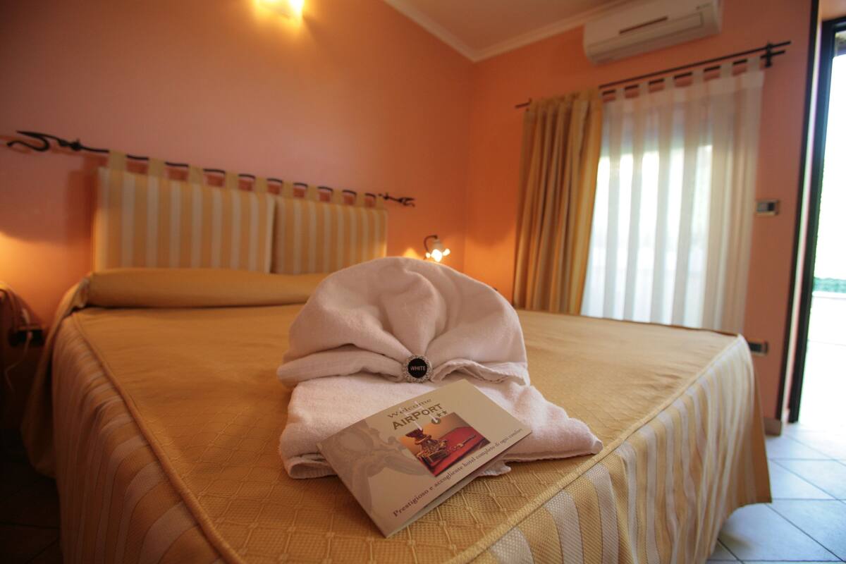 A hotel room with a bed, a window with curtains, an air conditioning unit, and a towel with a brochure on it.
