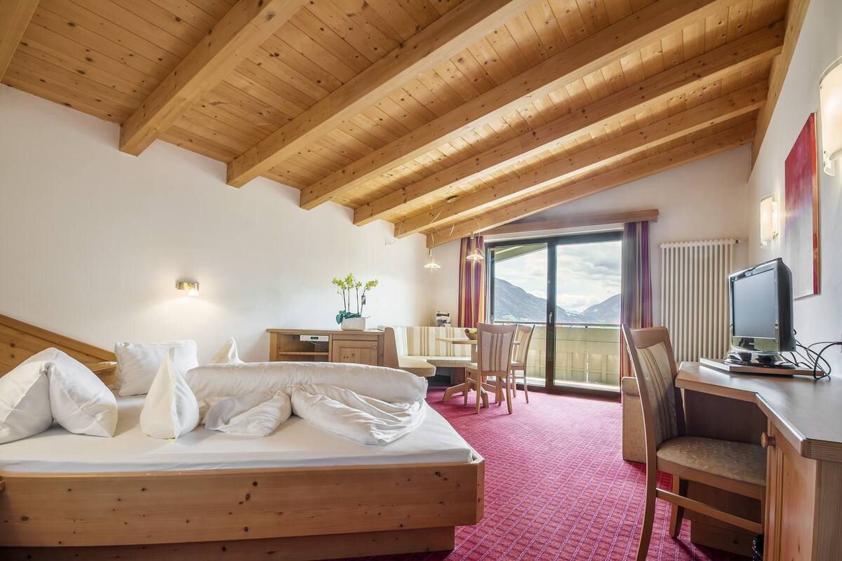A hotel room with a wooden ceiling, a bed, a desk, a chair, a television, and a view of mountains through a large window.