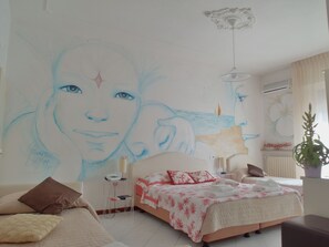 A bedroom with a large wall mural, a bed with a red and white floral bedspread, a bedside table with a lamp, and a potted plant. - Hotel Julia (Comacchio)