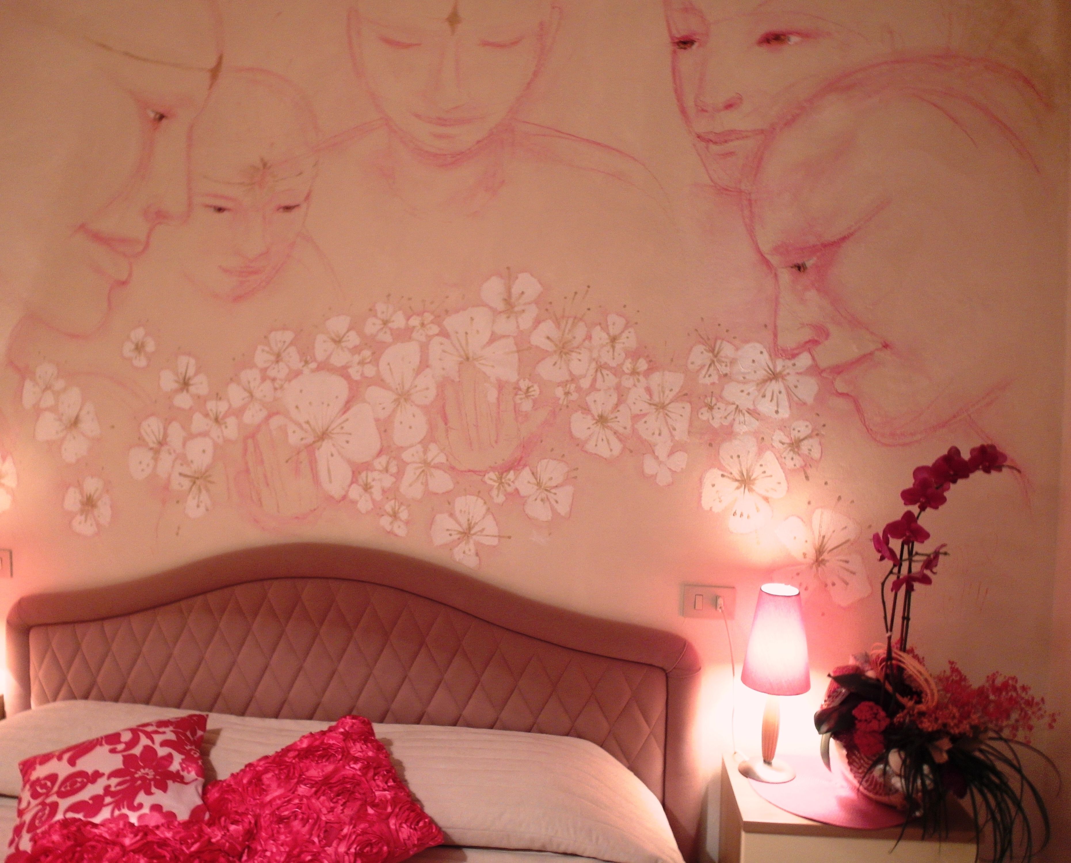 A bedroom with a bed, pillows, a nightstand with a lamp, and a wall with floral artwork.