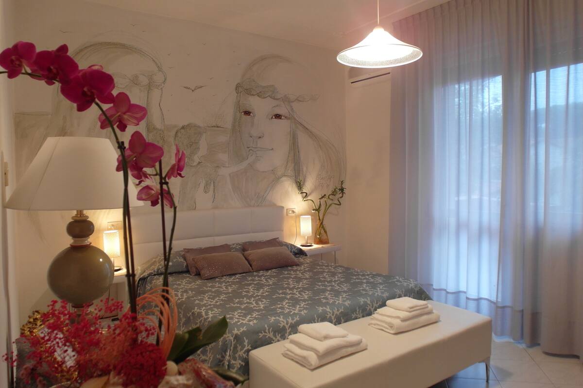 A bedroom with a bed, bedside lamps, a bedside table with a vase, and a large wall mural.