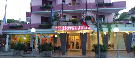 A hotel named 'Hotel Julia' with a modern facade, balconies with plants, and flags.