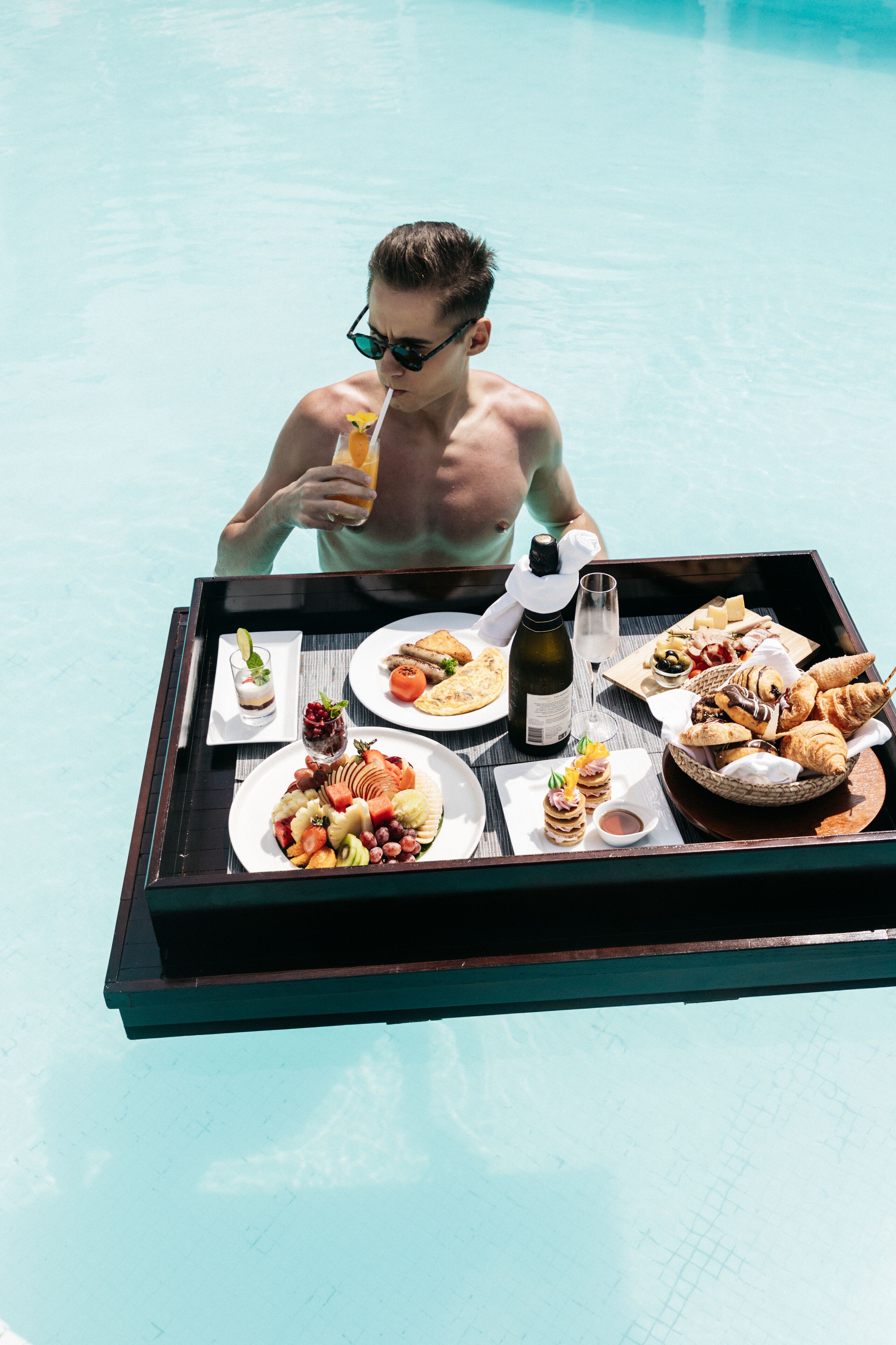 A dining table with a variety of food and drinks, including a bottle of wine, placed on a poolside deck.