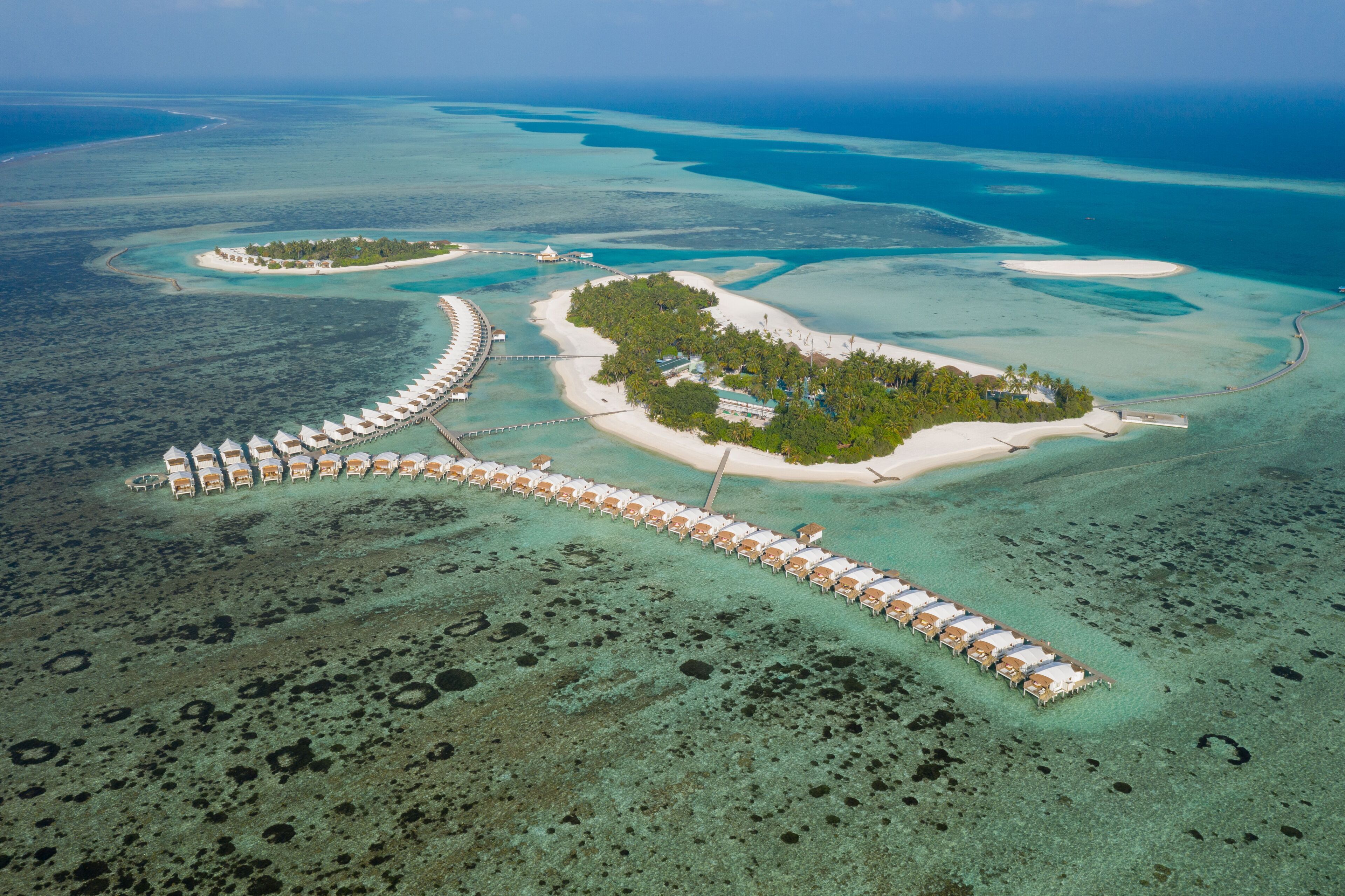 Vista aérea de un resort con una hilera larga de bungalós sobre el agua, conectados por un puente, rodeado de aguas turquesas cristalinas y pequeñas islas.
