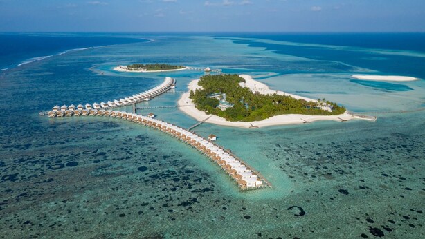 An aerial view of a resort with overwater bungalows connected by a long pier, surrounded by clear turquoise waters and small islands. - Cinnamon Hakuraa Huraa Maldives - All Inclusive Adults-Only (Hakuraa Huraa)