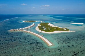 An aerial view of a resort with overwater bungalows connected by a long pier, surrounded by clear turquoise waters and small islands. - Cinnamon Hakuraa Huraa Maldives - All Inclusive Adults-Only (Hakuraa Huraa)