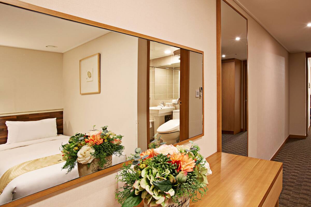 Una habitación de hotel con cama, un espejo grande, un baño visible a través del espejo y un escritorio de madera con un arreglo floral.