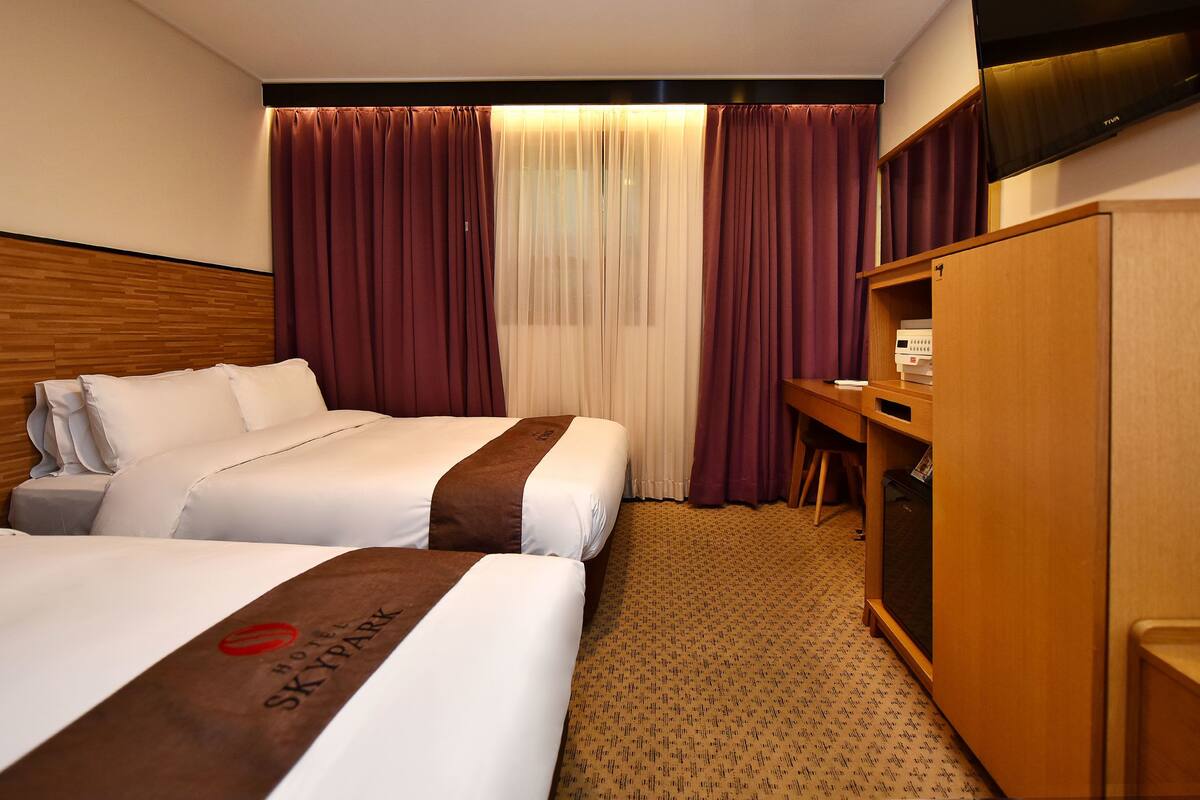 Habitación de hotel con dos camas, un cabecero de madera, un escritorio con silla, un armario y un televisor en la pared.