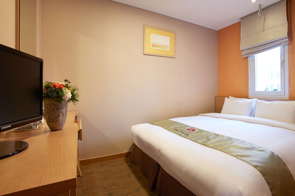A hotel room with a bed, a television on a desk, a window with a view, and a framed picture on the wall.
