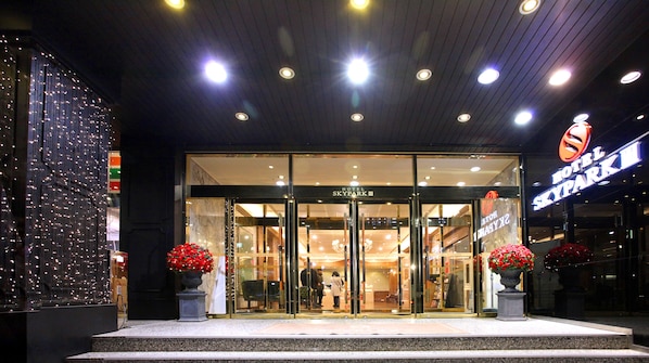 The entrance of Hotel Skypark III with glass doors, a sign above the entrance, and two potted plants with red flowers.