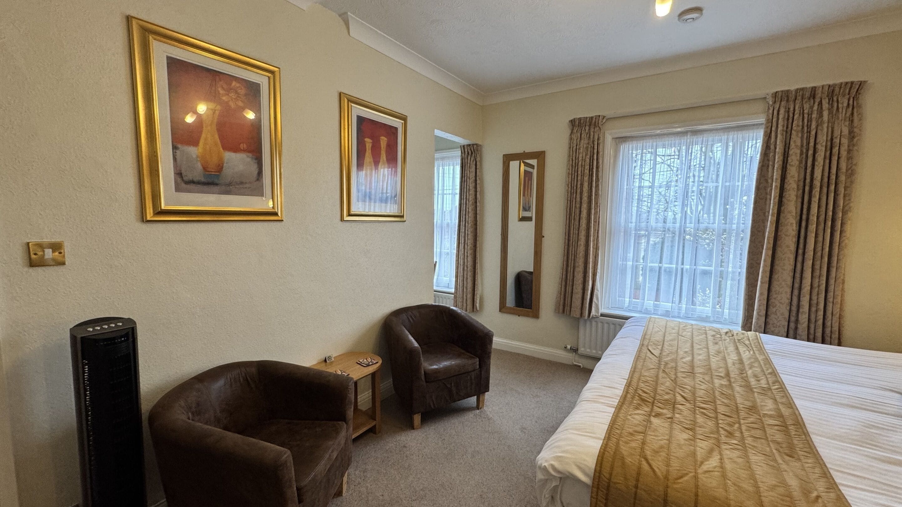Hotelkamer met een bed, twee stoelen, een tafeltje, een spiegel en twee ingelijste foto's aan de muur.