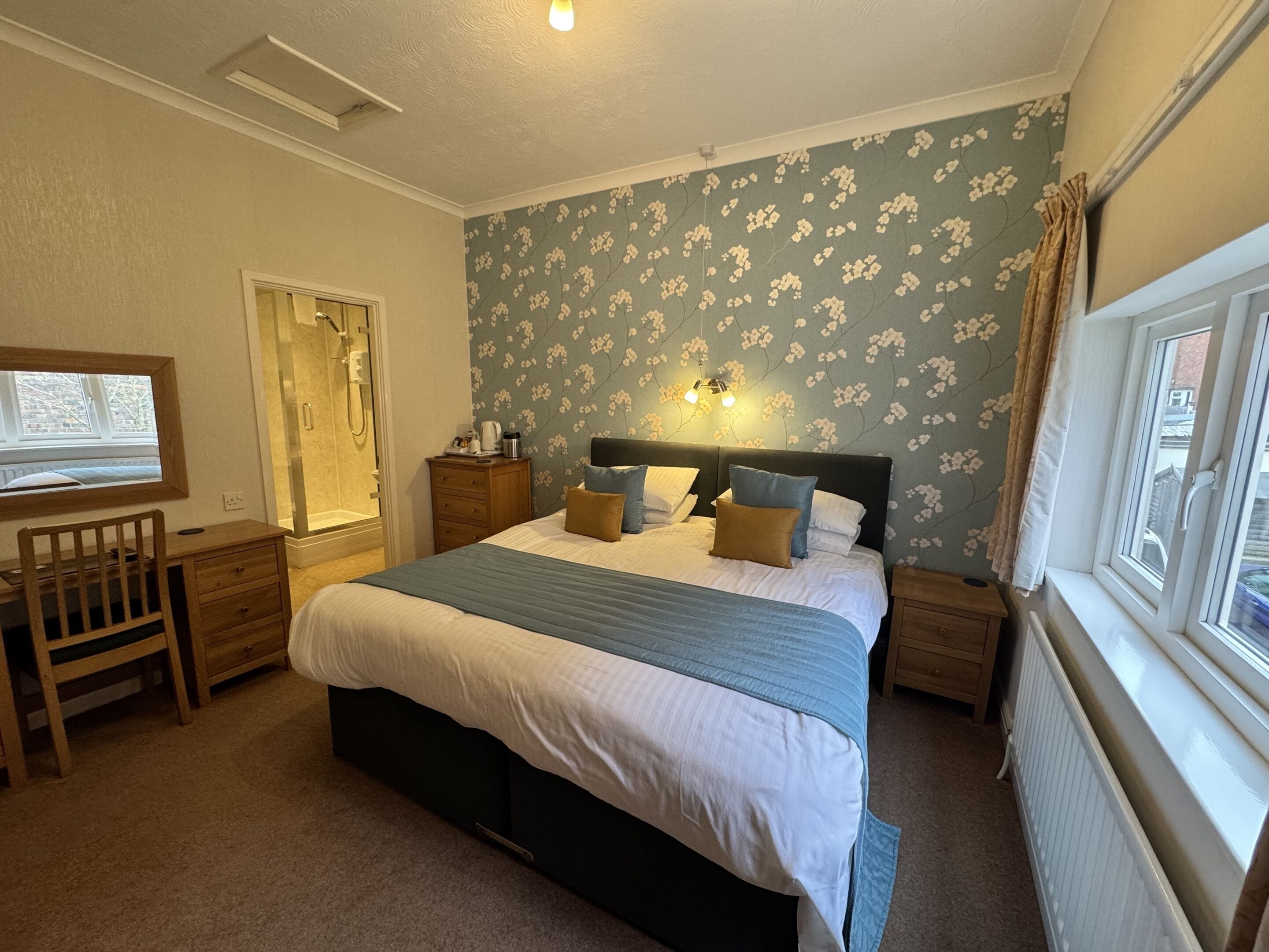 A bedroom with a bed, a desk, a chair, a nightstand, a dresser, a window, and a shower.