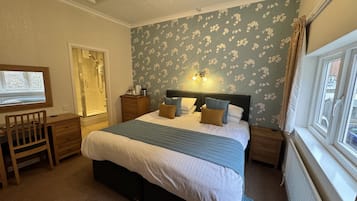 A bedroom with a bed, a desk, a chair, a nightstand, a dresser, a window, and a shower.